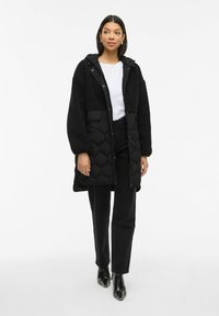 Sieviete, tērpta garā melnā polsterētā un flīsa mētelī, baltā blūzē, melnos biksēs un melnās potīšu zābakos, stāv uz balta fona.