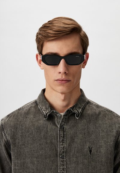 Sunglasses with a matte black frame, angular design, and dark lenses. Worn with a grey denim button-up shirt featuring a small black logo.