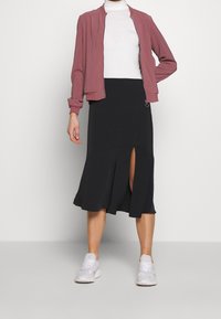 Blouson aviateur rose pâle zippé sur un col roulé blanc, associé à une jupe midi noire avec une fente à l'avant et des baskets blanches longueur cheville.