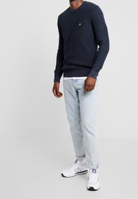 Marineblauer gerippter Strickpullover mit rundem Ausschnitt, kombiniert mit hell gewaschenen, geraden Jeans und weißen New Balance Sneakern. Kleines gelbes Logosymbol.