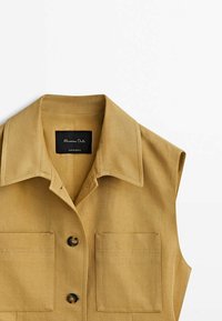 Beige ärmlös skjorta med krage i texturerad tyg. Har två framfickor och svarta knappstängningar. Märkesetikett synlig inuti.