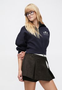 Navy zip-front sweatshirt with wide sleeves and embroidered logo; brown pleated skirt with textured fabric and side belt detail.