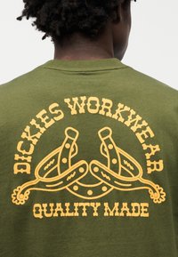 Ελιάς πράσινο μπλουζάκι με έντονο κίτρινο γραφικό κείμενο και σχέδιο στην πλάτη, που παρουσιάζει θέμα εργασιακής ένδυσης και κυκλικά μοτίβα.