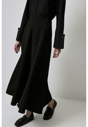 Femme portant une longue jupe noire, une chemise noire à manches longues avec des poignets boutonnés et des mocassins noirs brillants, debout sur un fond uni clair.