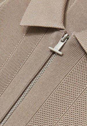 Beżowa, teksturowana tkanina swetra z zamkiem błyskawicznym z przodu. Posiada srebrny zamek oraz spiczasty kołnierz, eksponując szczegół o żebrowanym wzorze.