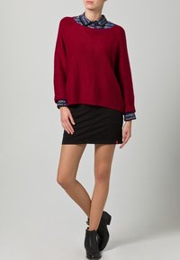 Burgundin neulepaita, joka on kerrostettu kuviollisen sinisen kauluspaita päällä, yhdistetty mustiin farkkuhameeseen ja mustiin nilkkureihin.