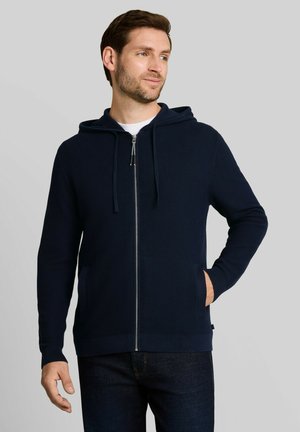 Homme aux cheveux bruns courts portant un sweat à capuche bleu marine zippé, la main gauche dans la poche et regardant vers la droite.