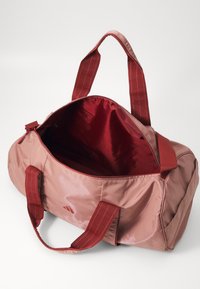 Sac de sport en nylon rose avec une fermeture éclair, doublure intérieure rouge et poignées en tissu rayé. Compartiment principal spacieux sans poches extérieures visibles.