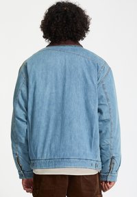 Lichtblauwe denim jacket met een bruine ribfluwelen kraag, ruime pasvorm, schoudernaden en drukknoopsluitingen op de mouwen. Eenvoudige achterkant zonder versieringen.