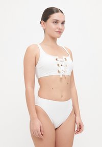 Wit bikini-set met een sportieve crop top met vetersluiting en gouden ogen, gecombineerd met hoge cut bikinibroekjes. Glad, rekbaar materiaal.
