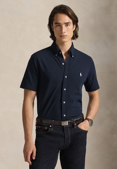 Σκούρο μπλε πουκάμισο με κοντά μανίκια και γιακά, λευκό λογότυπο στο στήθος, συνδυασμένο με σκούρα τζιν και καφέ ζώνη, μινιμαλιστικός σχεδιασμός.