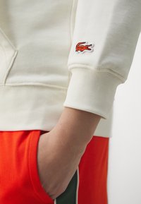 Felpa in cotone off-white con polsini a coste. Presenta un piccolo logo ricamato di un coccodrillo rosso e arancione sulla manica.