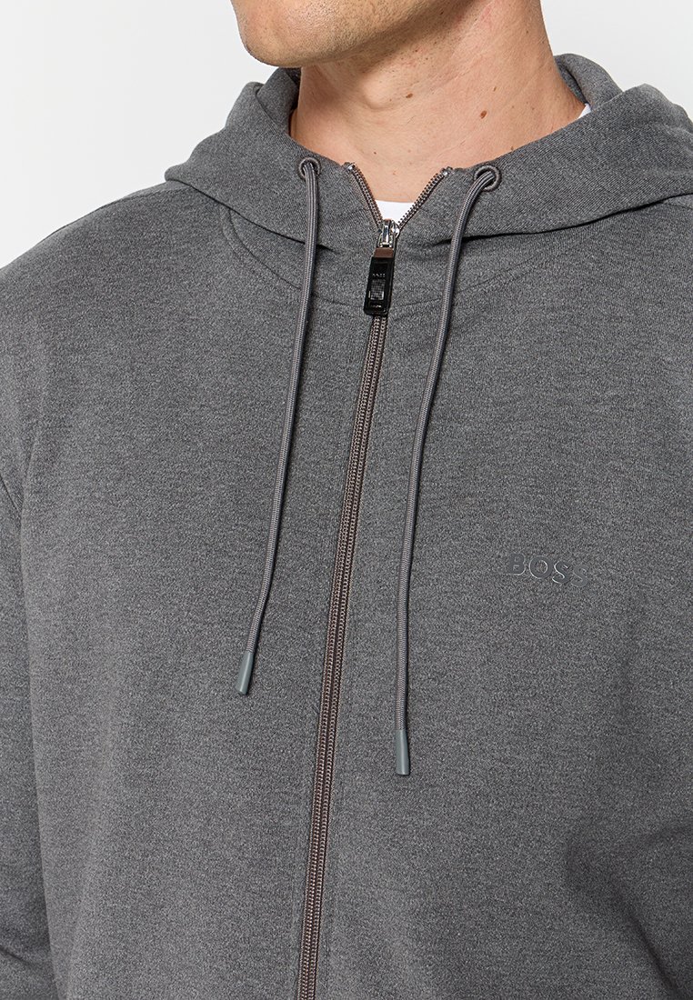 Man in een grijze ritssluiting hoodie met trekkoorden en een klein "BOSS" logo op de borst, gedeeltelijk zichtbare nek en kaak.