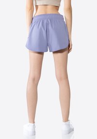 Shorts de sport lilas avec une taille élastique, poches latérales et une coupe ample, associés à des baskets blanches sur un fond neutre.