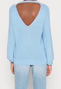 Jasnoniebieski sweter z dzianiny z dużym rozcięciem na plecach, długimi rękawami i ściągaczem u dołu. W zestawieniu z jasnoniebieskimi dżinsami w luźnym kroju.