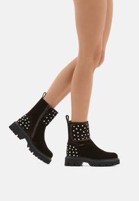 Stivali alla caviglia in suede marrone con suola in gomma spessa, caratterizzati da borchie bianche decorative ai lati e una cerniera laterale per una facile calzata.