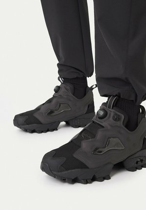 Scarpe da ginnastica Reebok Instapump Fury Trail nere e grigio scuro indossate con pantaloni e calzini neri, su sfondo bianco.
