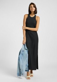 Abito maxi nero a coste con design senza maniche e spacco laterale, abbinato a un giubbotto di denim chiaro. Le calzature presentano un design tortuoso beige.