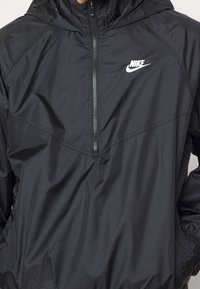 Mustavilla Nike-tuulitakki, jossa on vetoketjullinen etuosa, huppu ja logo. Ominaisuuksiin kuuluu kevyt, sileä tekstuuri ja kontrastiväriset ompeleiden yksityiskohdat.