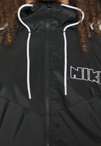 Svart vindjacka i lätt material med heldragkedja, vita snören och broderad "NIKE"-logga på bröstet.