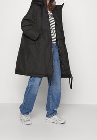 Černý dlouhý péřový kabát s kapucí, zipovým zapínáním a dvěma postranními kapsami, v kombinaci s pruhovaným tričkem a modrými džíny. Obuv: světlejší tenisky.