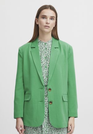 Blazer verde su misura con rever a punte, chiusura a un bottone e due tasche anteriori. Indossato sopra un vestito a righe con colletto.