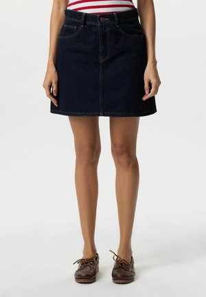 Kobieta w ciemnoniebieskiej dżinsowej mini spódniczce z przednimi kieszeniami i brązowych mokasynach, stojąca na jednolitym białym tle.