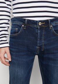 Jeans de mezclilla azul oscuro con áreas descoloridas prominentes, diseño clásico de cinco bolsillos, cierre de botón de latón y costuras en contraste. Camisa de manga larga a rayas visible.