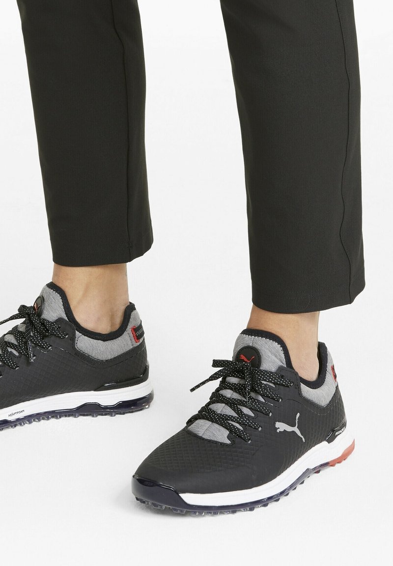 Baskets Puma noires et grises avec semelles blanches portées avec un pantalon noir, visibles des chevilles vers le bas sur fond blanc.