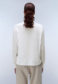 Kermetin värinen pitkähihainen paita, jossa on rento istuvuus, sileä tekstuuri, pyöreä pääntie ja yksityiskohtia alaosassa, käytetään beigen väristen housujen kanssa.