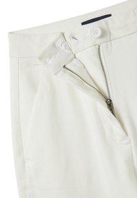 Pantaloni bianchi con una zip parzialmente aperta, chiusura con bottone e passante interno con bottone sulla vita.