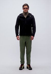 Homme portant une veste noire, un pantalon vert olive, des chaussures de randonnée et des lunettes de soleil, debout devant un fond blanc.