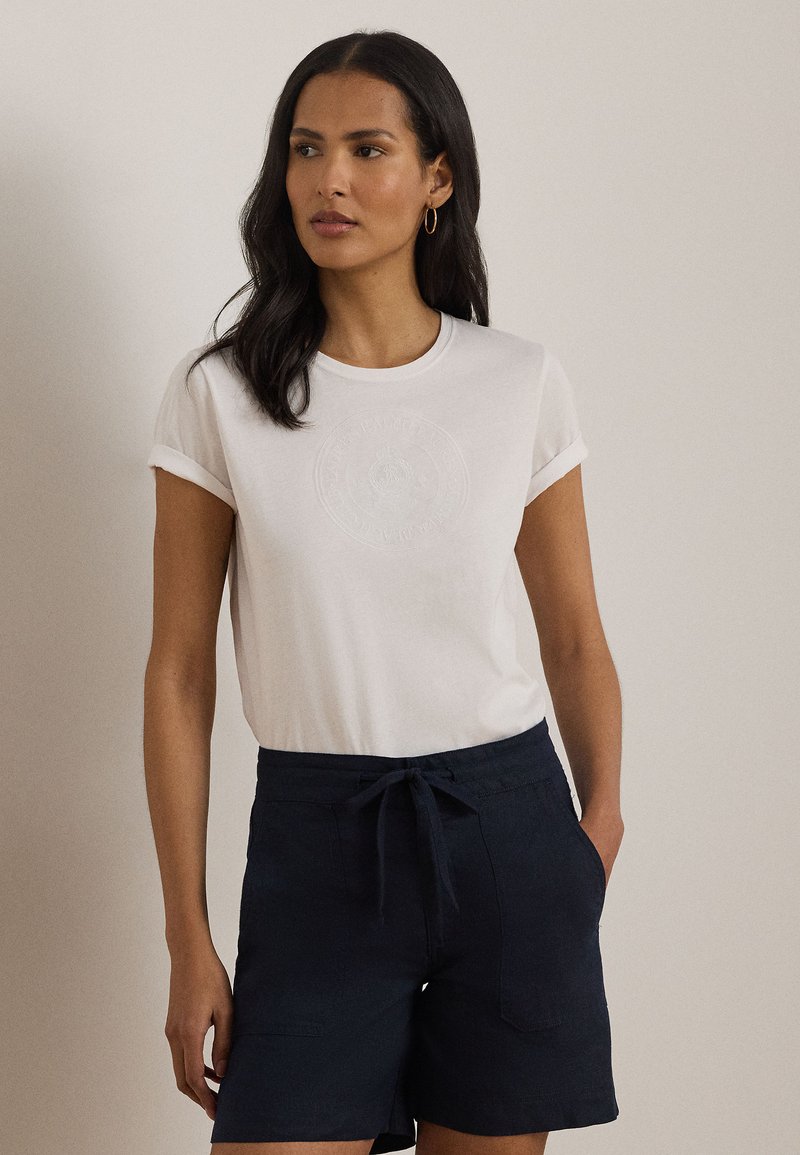 Γυναίκα με μακριά σκούρα μαλλιά, φοράει λευκό t-shirt με ρολερ μανίκια και σκούρα μπλε σορτς με κορδόνι, στέκεται με το ένα χέρι στην τσέπη.