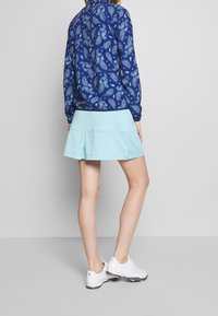 Modrá košile s paisley vzorem a dlouhým rukávem, doplněná světle modrou skládací sukní. Modelka má na sobě bílé sportovní boty.