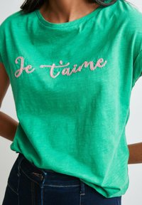 Camiseta de algodón verde con texto bordado en rosa "Je t'aime", con un corte relajado y cuello redondo. Textura suave, mangas cortas.