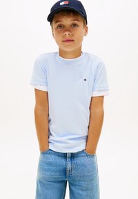 Αγόρι που φοράει ανοιχτό μπλε μπλουζάκι Tommy Hilfiger, μπλε τζιν και μπλε σκουφάκι με κεντημένο λογότυπο, στέκεται με τα χέρια στις τσέπες μπροστά σε λευκό φόντο.