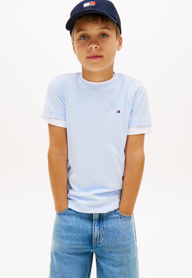 Αγόρι που φοράει ανοιχτό μπλε μπλουζάκι Tommy Hilfiger, μπλε τζιν και μπλε σκουφάκι με κεντημένο λογότυπο, στέκεται με τα χέρια στις τσέπες μπροστά σε λευκό φόντο.