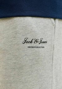 Graue Jogginghose mit schwarzem "Jack & Jones Archive Selection" Logo in der Nähe der Tasche, kombiniert mit einem dunkelblauen gerippten Bund des Pullovers.