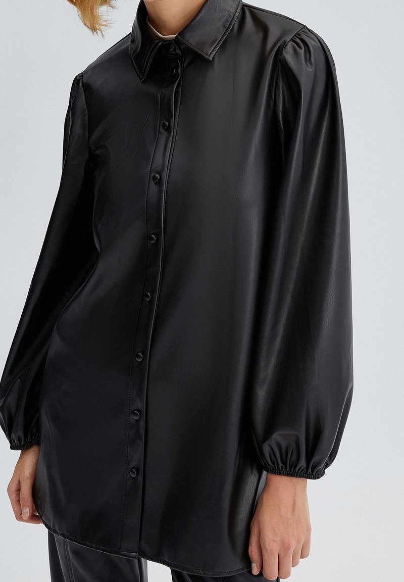 Svart konstläder skjorta med lång ärm och knäppning, puffärmar och elastiska manschetter, bärs av en person med kort hår.