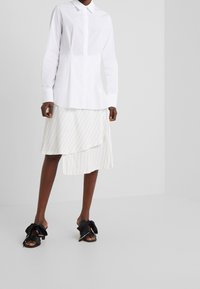Chemise blanche boutonnée à manches longues, associée à une jupe blanche asymétrique rayée. Sandales noires ornées de nœuds oversize.