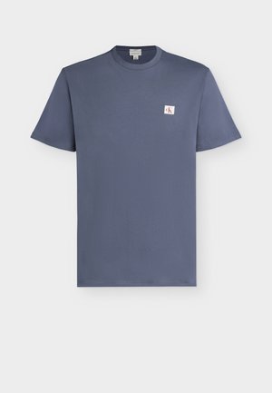 T-shirt bleu à manches courtes avec col rond, avec un petit patch logo carré blanc comportant les lettres rouges "cK" sur la poitrine gauche.