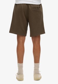 Pantalones cortos marrones de corte relajado, con detalles de bolsillos laterales y dobladillo cosido. Combinados con calcetines claros y zapatos casuales, mostrando un diseño sencillo.