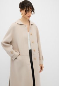 Cappotto in lana beige chiaro con colletto, sei bottoni in legno, tasche laterali e maniche larghe. Texture liscia, forma sagomata.