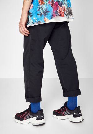 Personne portant un pantalon noir retroussé, des chaussettes bleu vif, des baskets Adidas noires avec des détails rouges, et une chemise florale colorée.