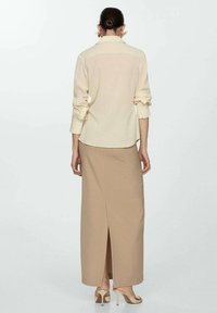 Blusa de color beige claro con mangas remangadas y un cuello, combinada con una falda beige larga hasta el suelo que presenta una abertura en la parte trasera. Diseño minimalista.