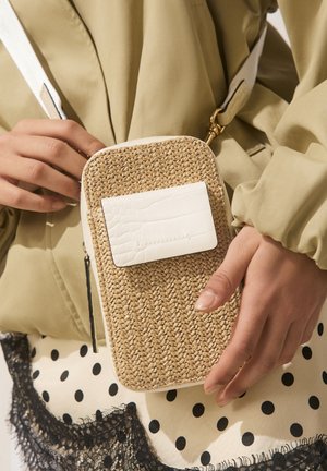 Mâini ce țin o geantă mică țesută din paie, cu o pată din piele albă, purtată peste o jachetă bej și o fustă cu buline.