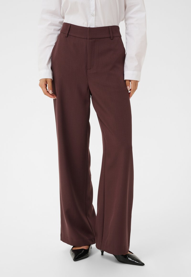Personne portant un pantalon large marron taille haute, une chemise blanche boutonnée et des talons noirs pointus, debout sur un fond uni.
