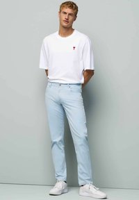 Lichtblauwe slim fit broek met een rits en knoopsluiting aan de voorkant, gecombineerd met een wit t-shirt met korte mouwen en een rood logo.