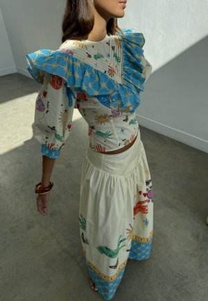 Femme portant une robe à motifs colorés avec un col à volants bleu et des manches bouffantes, debout dans une pièce ensoleillée avec un sol gris.