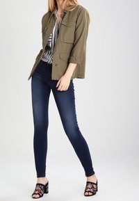 Mujer que lleva una chaqueta verde de botones sobre una camiseta de rayas en blanco y negro, jeans ajustados azul oscuro y sandalias de tacón negras con bordado floral.